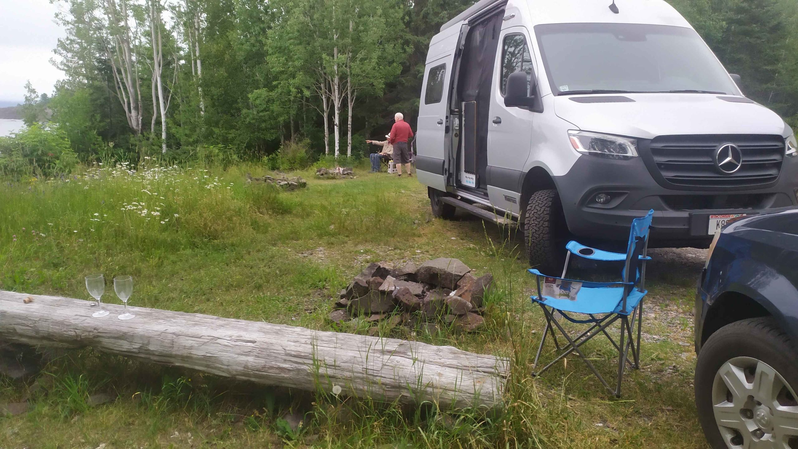 un van avec une porte ouverte ainsi qu'une chaise de camping dépliée Deux coupes à champagne en plastique sont déposés sur un rondin de bois