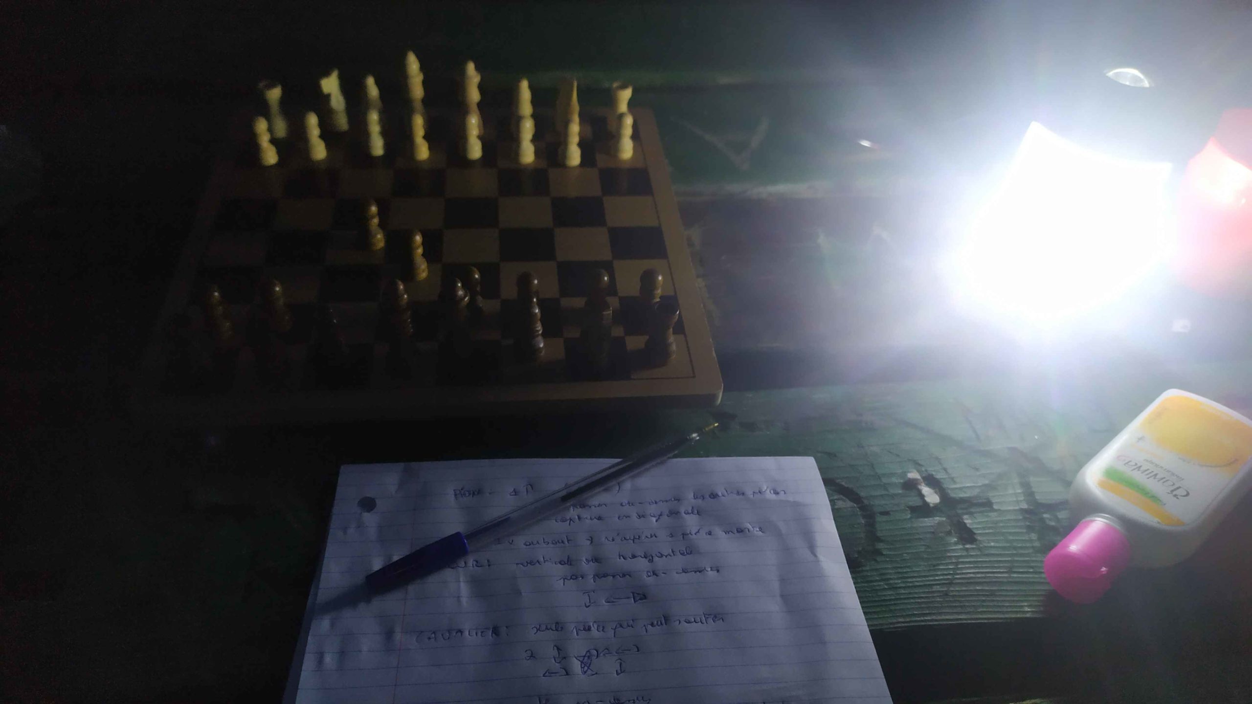 Un plateau d'échecs éclairé par une petite lampe d'extérieur avec un papier où sont notés les règles 